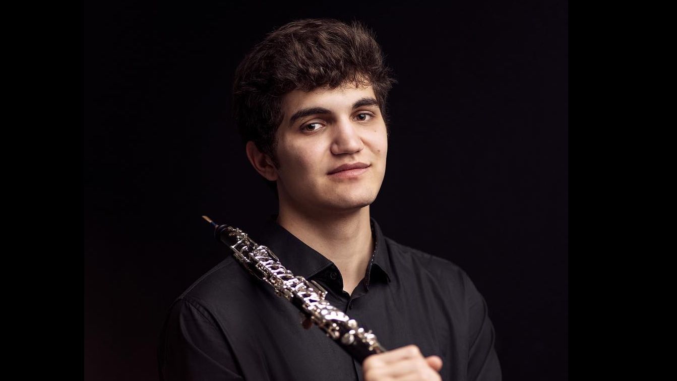 Junger Mann mit dunklen Locken und Oboe in der Hand vor dunklem Hintergrund